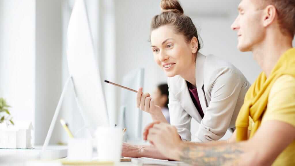 Two colleagues working at a desk with computers. A woman wearing a white blazer, showcasing leader characteristics, is pointing at a screen with a pencil and smiling, while a man with tattoos and a yellow shirt looks on attentively. The background shows a bright, modern office space.