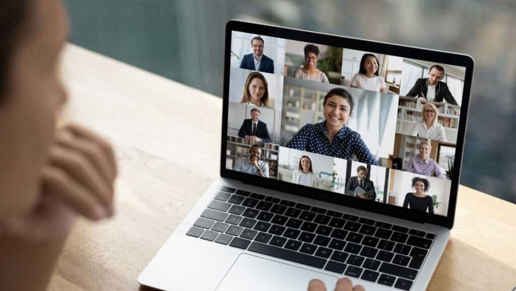 A person sits at a desk, engaging in a video conference on a laptop. The laptop screen displays ten participants, each in a separate window, discussing small business sustainability. The desk has a soft background blur.