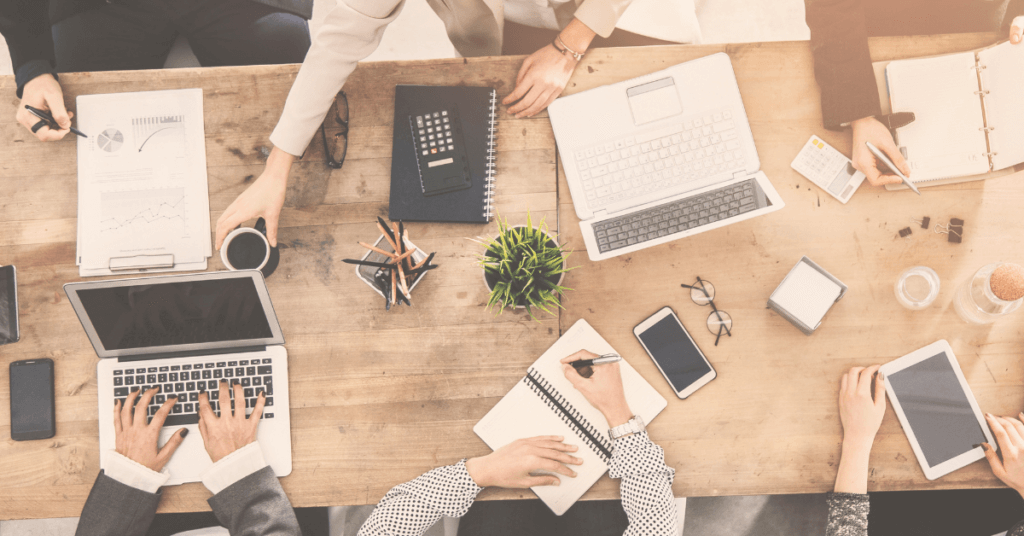 Top-down view of a wooden table with people working collaboratively. Laptops, notebooks, smartphones, coffee, documents, and office supplies are spread across the table. A small potted plant in the center adds a touch of greenery as they focus on continuing to diversify and grow in 2023.