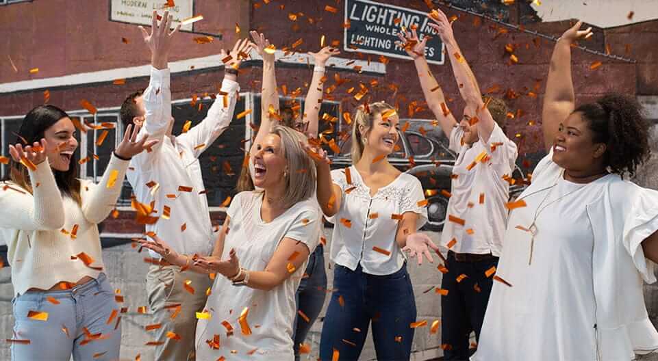 A group of six people, dressed casually in light-colored clothing, celebrate joyfully indoors with orange confetti falling around them. They are smiling, raising their arms, and appear to be having fun. The background shows a partial mural and industrial decor.