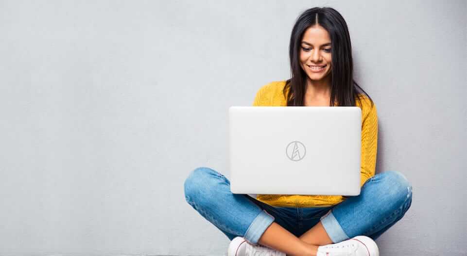 A person with long dark hair sits cross-legged on the floor against a gray wall, smiling while using a laptop. They are wearing a yellow sweater, blue jeans, and white sneakers. The laptop has a simple logo on the back.