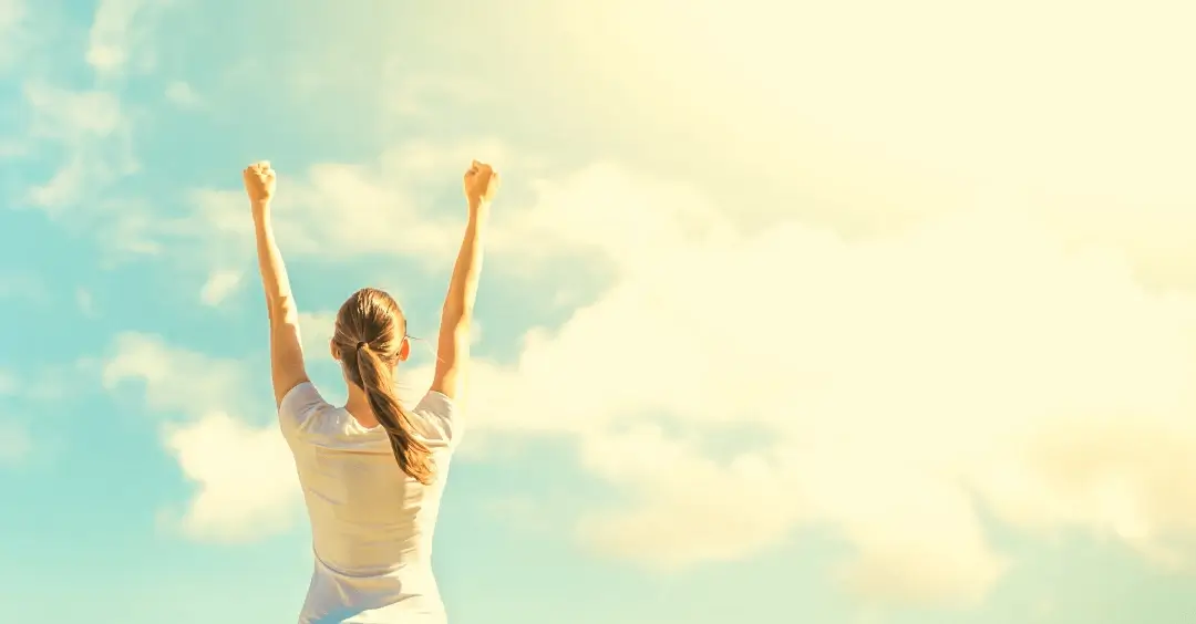 A woman stands with arms raised against a bright sky, symbolizing personal achievement, freedom, and a sense of triumph.