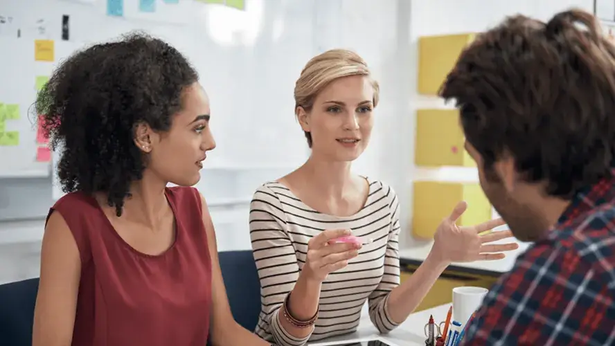 Three individuals engage in a discussion around a table, surrounded by colorful sticky notes on a wall, fostering a collaborative atmosphere.