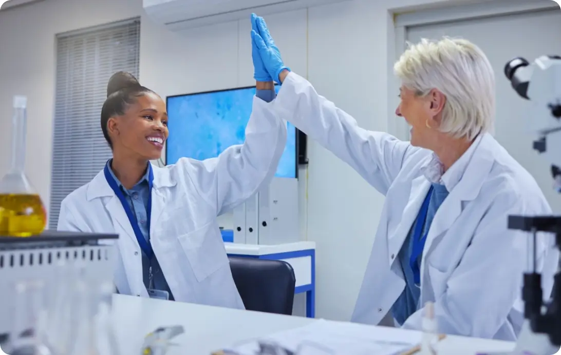 Two scientists in lab coats and gloves high-five in a modern laboratory, celebrating a successful experiment amid scientific equipment and glassware.