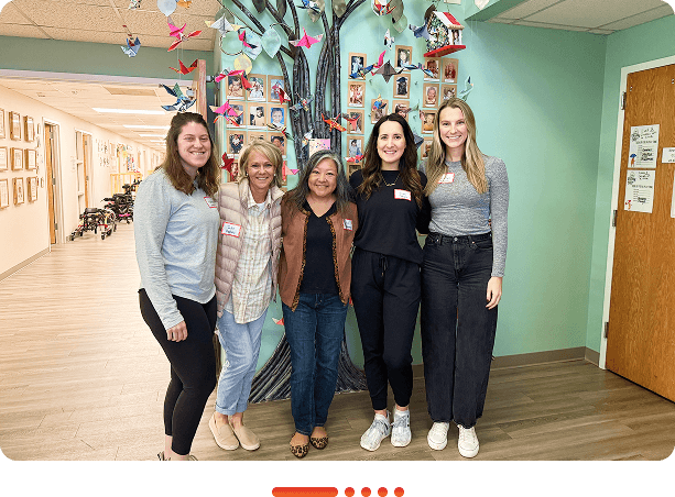 Five women standing together in front of a colorful mural of a tree decorated with photos and origami, symbolizing unity, creativity, and community in a positive work environment.