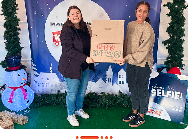 Two women are holding a festive box labeled "Happy Holidays!" in front of a winter-themed backdrop with a snowman.