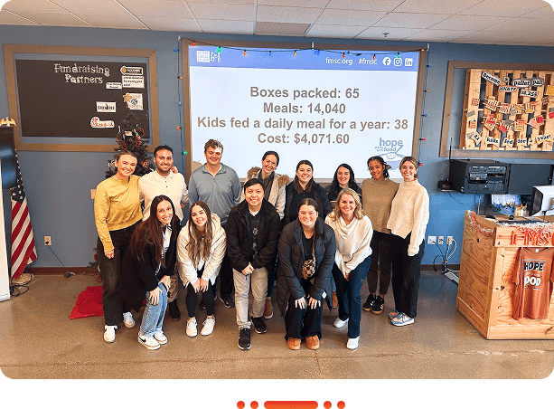 A group of diverse individuals poses in front of a screen displaying donation stats for a meal packing initiative, highlighting their impact.