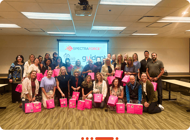 A diverse group of professionals poses together in a conference room, holding bright pink bags, with a presentation screen in the background.