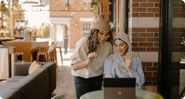 Two women sit at a table in a cozy café, one gesturing while the other waves, both engaged in a conversation over a laptop—highlighting a collaborative and friendly work environment.
