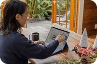 A person sits at a wooden table, using a tablet with a coffee cup and decorative items nearby, surrounded by greenery—creating a relaxed and productive work environment.