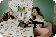 Two women sit at a stylish, colorful table, engaging in conversation and using a tablet in a modern, green-themed workspace—illustrating collaboration and innovation in a contemporary office setting.