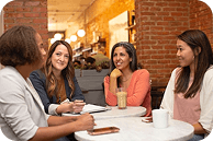 A group of women engage in conversation at a cozy cafe, seated around a table with drinks and notebooks—creating a warm and inviting atmosphere for collaboration and connection.
