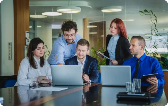 A group of professionals collaborate around a conference table with laptops in a modern office setting, representing teamwork and productivity.
