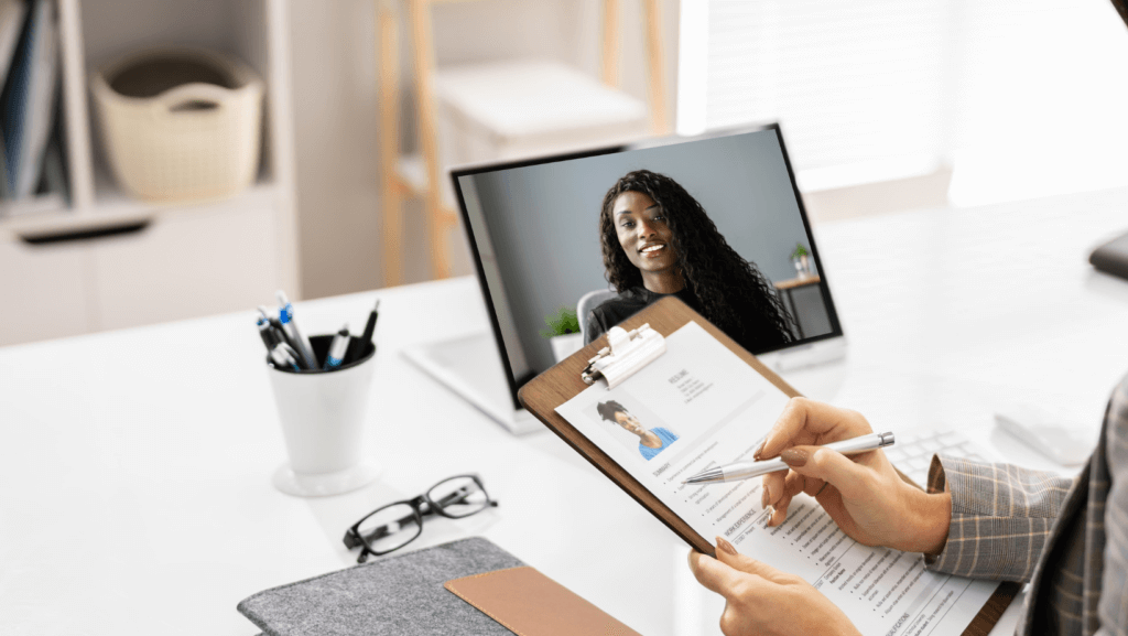 A person holds a clipboard with a printed resume while conducting a virtual interview on a laptop. The laptop screen shows a woman with long hair, smiling. A pen, glasses, and office supplies are on the desk.