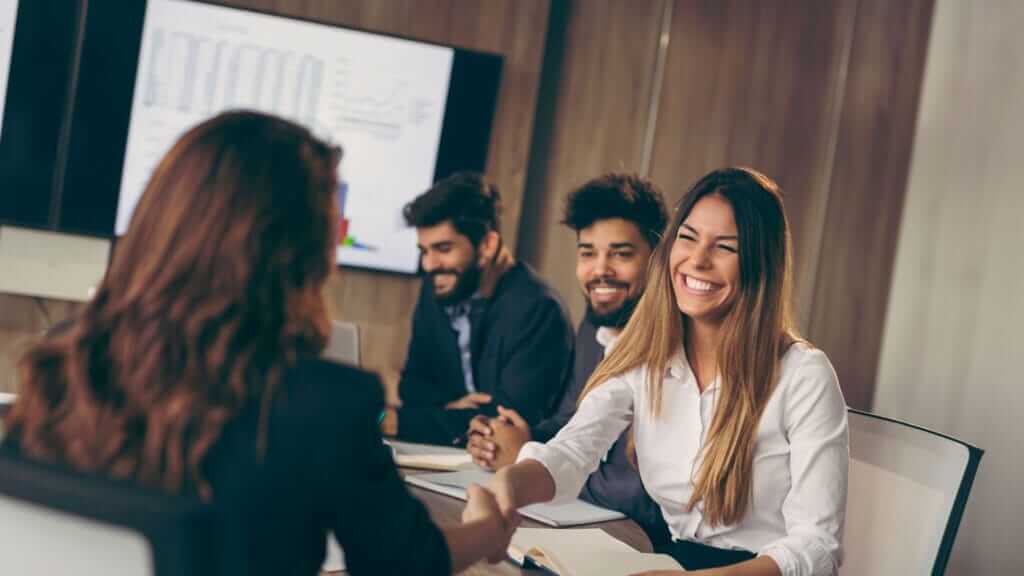 A group of people are seated at a conference table in a modern office. A smiling woman is shaking hands with another person, possibly concluding a meeting or reaching an agreement. Other colleagues are present, and a chart is displayed on a screen in the background.