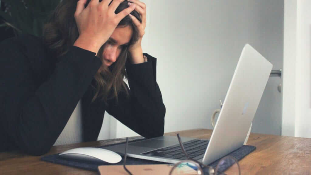 A person sits at a wooden table, looking stressed with their hands in their hair, facing a laptop. Glasses, a mouse, and a phone are also on the table. Amidst the struggle, it's easy to remember that even inspirational women have faced moments like these.