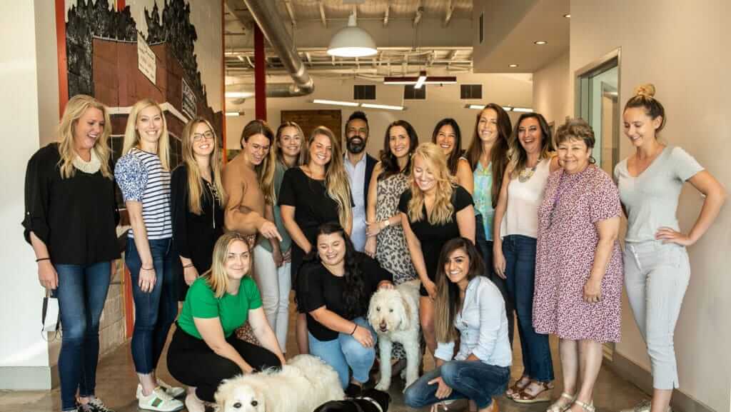 A group of sixteen women smiling and posing together indoors, with two large dogs in the front. The well-lit room, adorned with artwork and an industrial ceiling, serves as the backdrop for this joyous gathering. Several women are casually dressed, some kneeling or crouching near the dogs, perhaps after a day of engaging training courses.