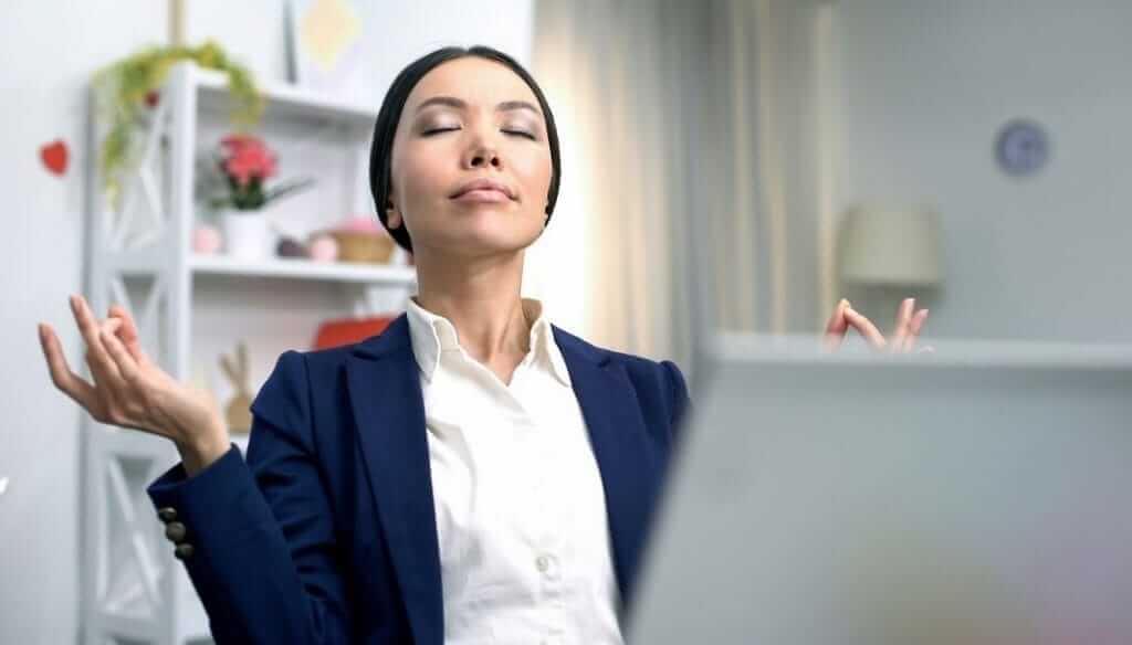 A person dressed in a blue blazer and white shirt is sitting at a desk with eyes closed and fingers forming a meditation pose. Behind them, there are shelves with decorative items and a window with a curtain, suggesting a calm indoor environment.