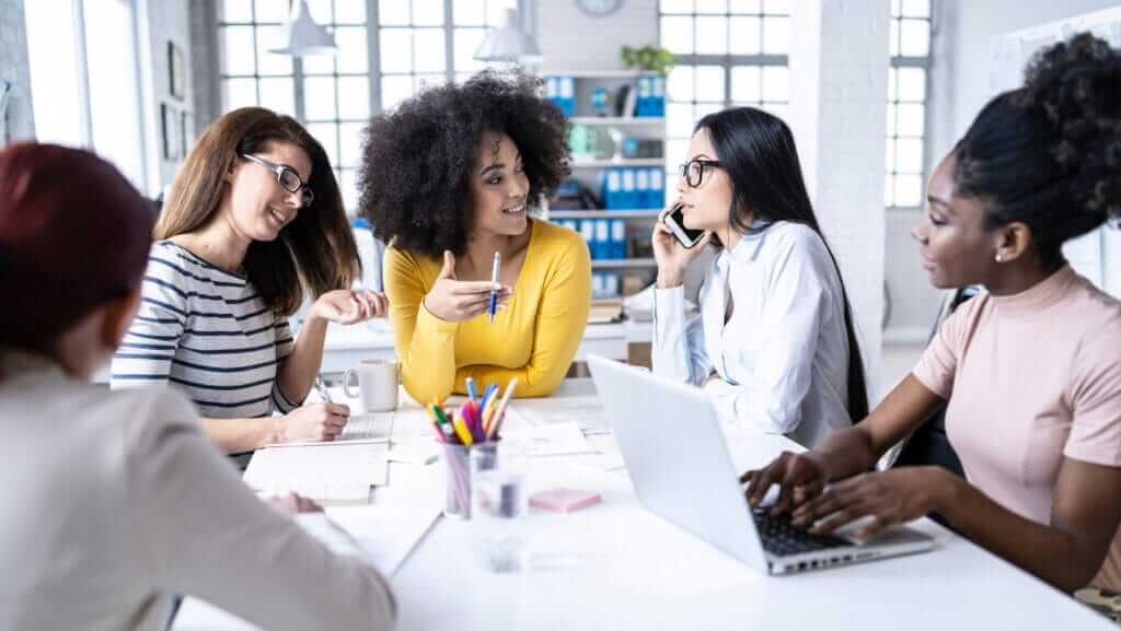 A diverse group of five inspirational women having a lively discussion in a bright, modern office. One woman speaks on the phone, another types on a laptop, while the others engage in conversation, surrounded by documents and office supplies on the table.