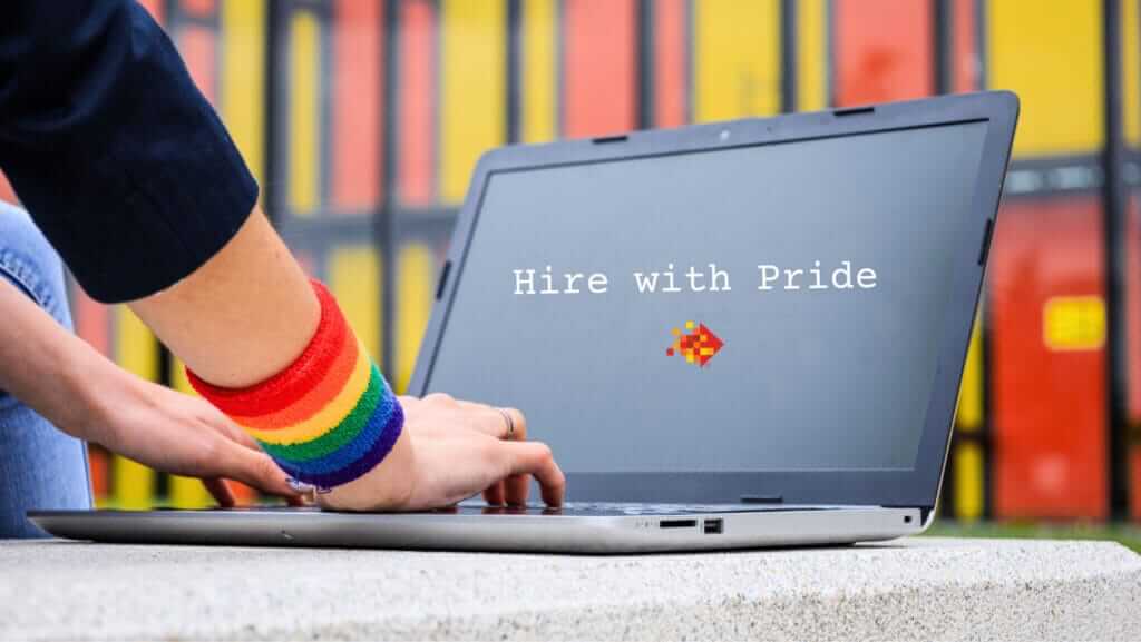 Close-up of a person wearing a rainbow wristband typing on a laptop. The screen displays the text "Hire with Pride" along with a pixelated rainbow arrow pointing to the right. The colorful, blurry background suggests an environment that values small business sustainability.