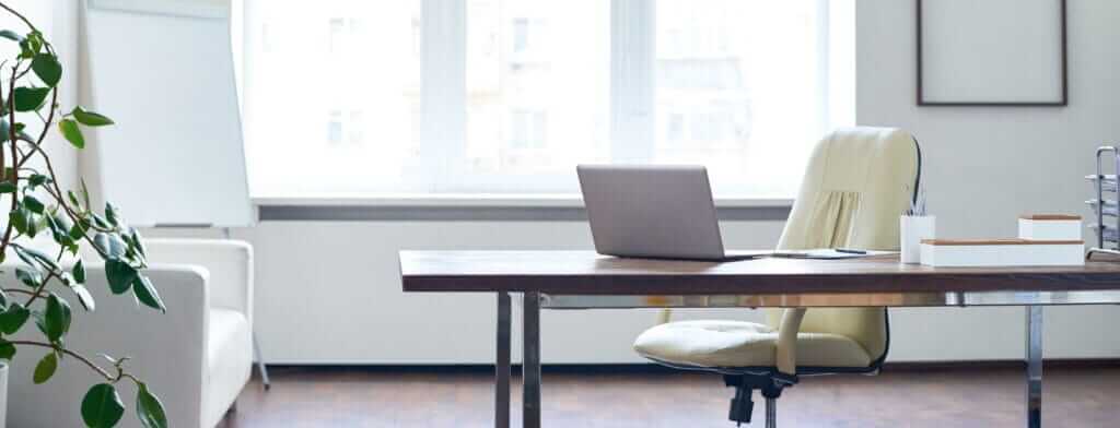 A modern, well-lit office space with a wooden desk, beige swivel chair, and open laptop ready for training courses. The desk also has a white organizer holding pens and notepads. A potted plant sits to the left, and large windows in the background let in ample natural light.