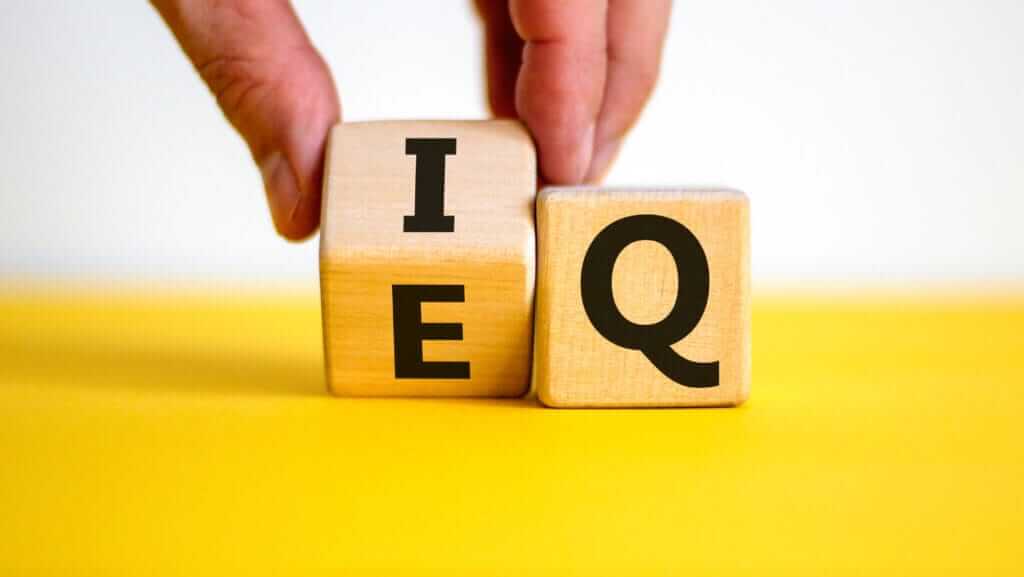 A close-up of two wooden blocks against a yellow background. One block has the letter "I" on the left side, and the letter "E" is on top. The other block has the letter "Q" on the front side. A hand is adjusting the position of these blocks, reminiscent of how inspirational women shape their paths.