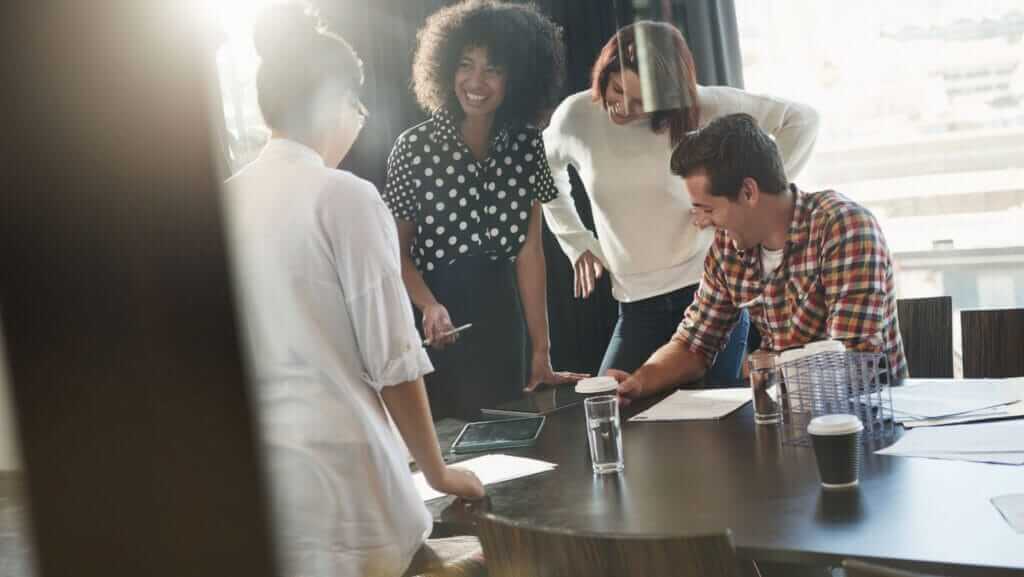 A group of four people, three women and one man, are having a lively discussion around a conference table with papers and coffee cups on it. They appear to be working collaboratively in a well-lit office environment with large windows, focused on small business sustainability initiatives.