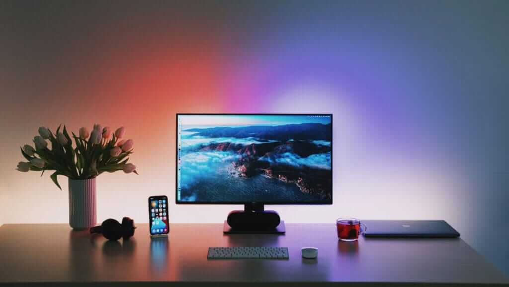 A minimalist desk setup features a widescreen monitor displaying a scenic landscape, surrounded by a laptop, smartphone, keyboard, headphones, and a cup of tea. A vase with tulips pays tribute to inspirational women. Ambient lighting in shades of red and blue adds a colorful backdrop to the scene.