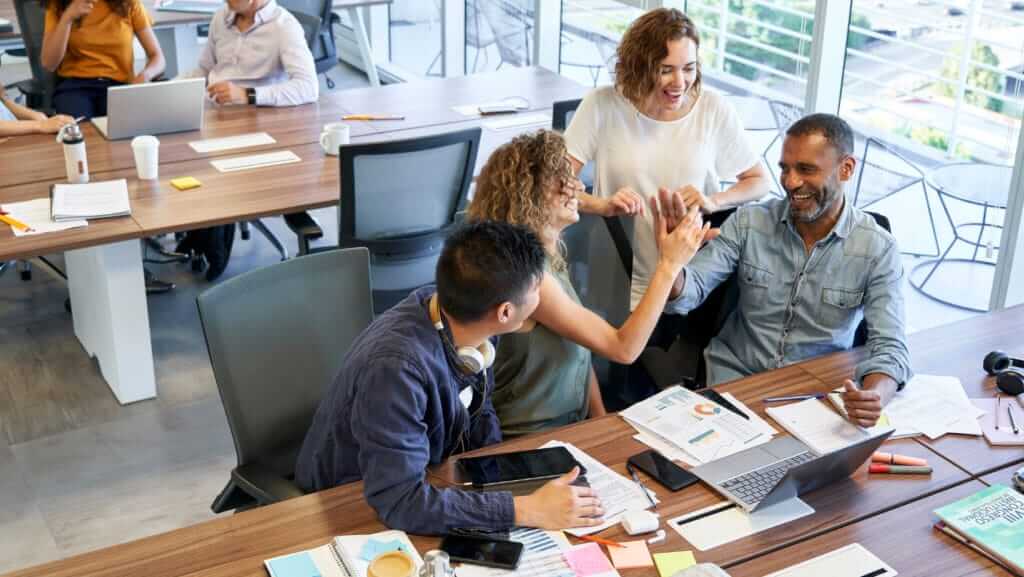 A diverse group of colleagues at a modern office table are engaged in lively conversation and teamwork, discussing recent training courses. One standing individual high-fives a seated colleague, while others work on laptops and tablets. The atmosphere is collaborative and energetic.