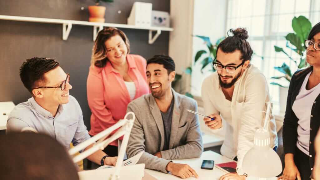 A diverse group of five people, three men and two women, are gathered around a desk in an office. Smiling and engaged in discussion, they exhibit good business traits. One man holds a pen, gesturing towards a piece of paper. Shelves, plants, and a window are in the background.