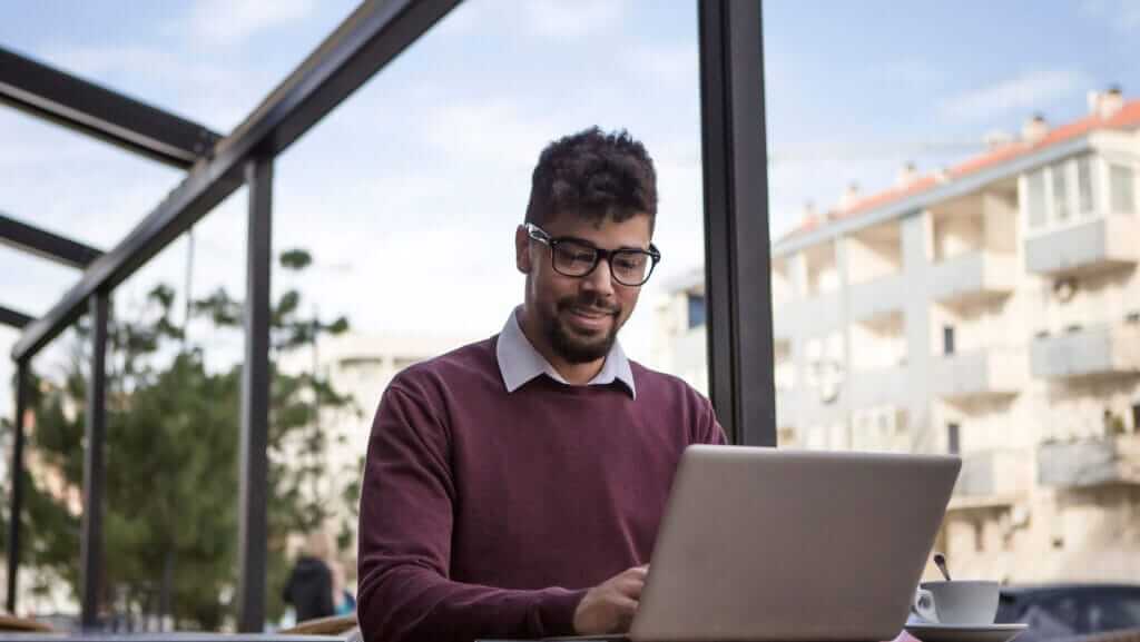 A person with glasses and a beard works on a laptop at an outdoor café. They are wearing a maroon sweater over a collared shirt. A cup of coffee is on the table beside them, next to a book about inspirational women. The background shows a clear sky and buildings.