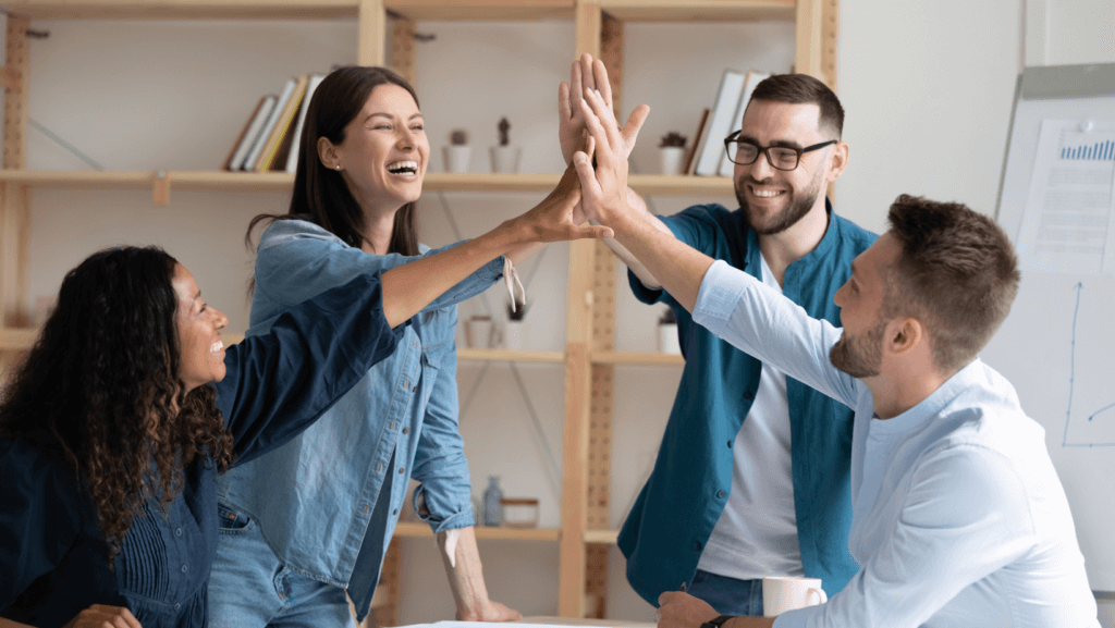 Four colleagues in casual attire smile and high-five each other in an office setting, with shelves and a whiteboard in the background. They appear to be celebrating or sharing a moment of success, showcasing excellent employee engagement.