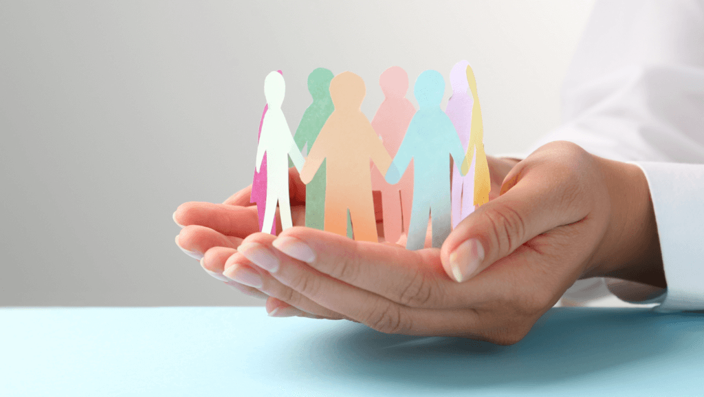 A person's hands gently cradle a colorful paper chain of figures, symbolizing unity and togetherness within a diverse and inclusive workforce. The figures are cut out of pastel-colored paper, and the background is softly lit, emphasizing the supportive gesture of the hands.