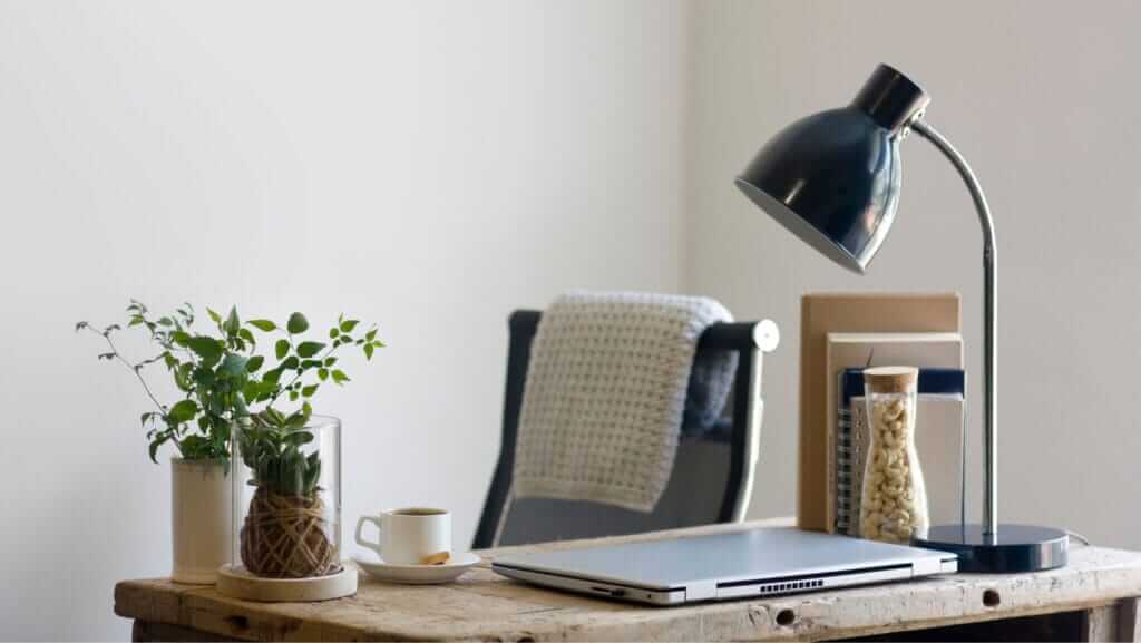 A minimalist workspace with a wooden desk holding a closed laptop, a small plant in a glass pot, a cup of espresso on a saucer, and a black desk lamp. In the background, showcasing leader characteristics of organization and calm efficiency, there's a chair with a blanket draped over it and two books standing upright.