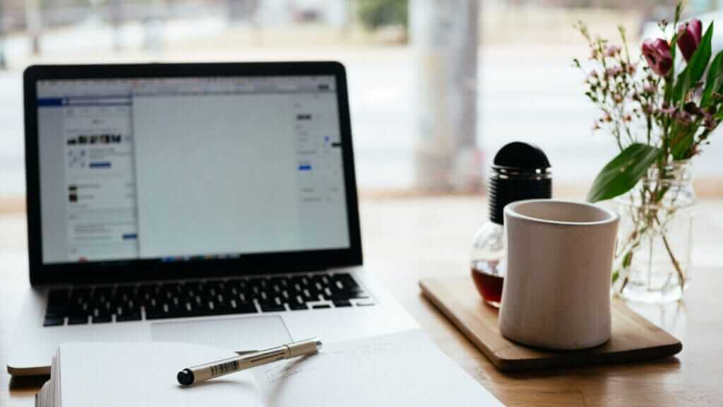 A laptop displaying a partially open document sits on a wooden table next to a notebook with a pen resting on it. To the right of the laptop, there is a white mug and a small glass jar with a black lid on a wooden tray, embodying leader characteristics in its organized and thoughtful setup, while a small vase with flowers completes the scene.