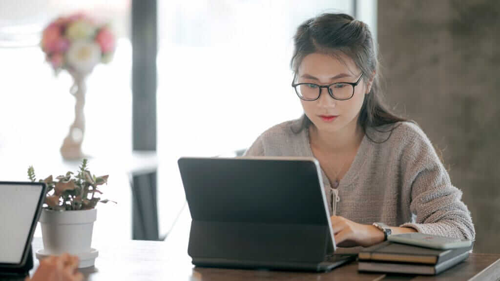 A woman with glasses, wearing a grey sweater, sits at a table working on a laptop. A notebook is beside her, and a potted plant is placed nearby. She is focused on her work in a well-lit, modern room with a blurred background, diligently researching training courses.