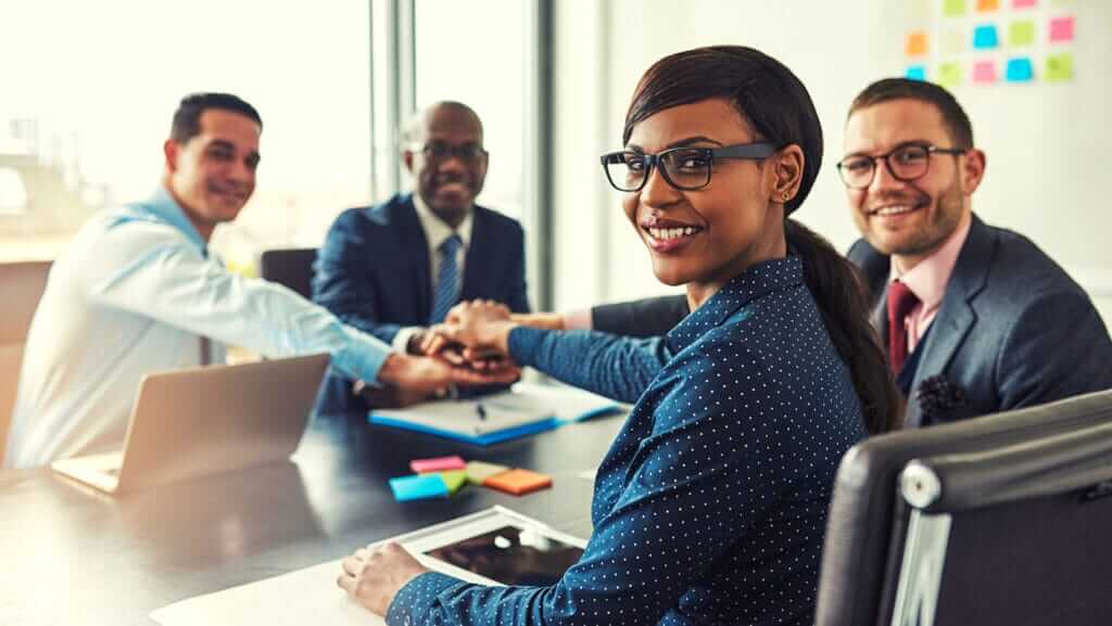 A group of four businesspeople sit around a table in a modern office. They are smiling and stacking their hands together in a gesture of teamwork. The woman in the foreground, an inspirational figure, wears glasses and a polka dot blouse. Laptops and colorful sticky notes are on the table.