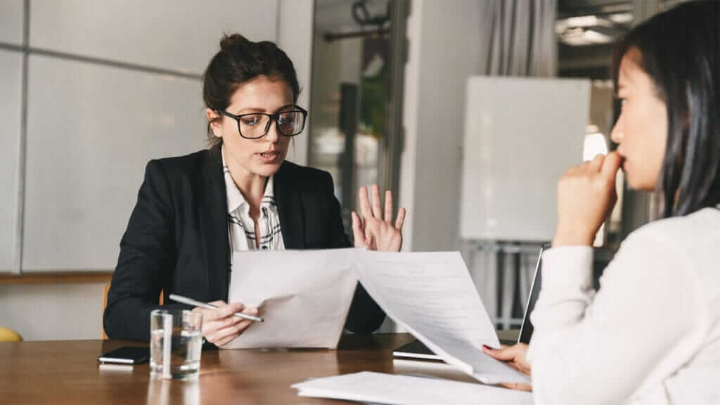 Two women are sitting across from each other at a table in a professional setting. The woman on the left, wearing glasses and a suit, is holding and discussing a document, while the woman on the right, with a pensive expression, listens attentively. A glass of water is on the table.