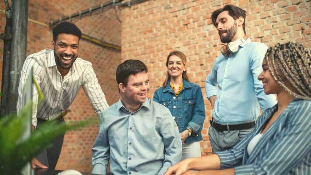 A group of five people are gathered in an office with brick walls, smiling and engaging in a lively conversation. Reflecting the spirit of inclusion, three are standing—two men and one woman—while another man and woman are seated. One standing man has headphones around his neck.