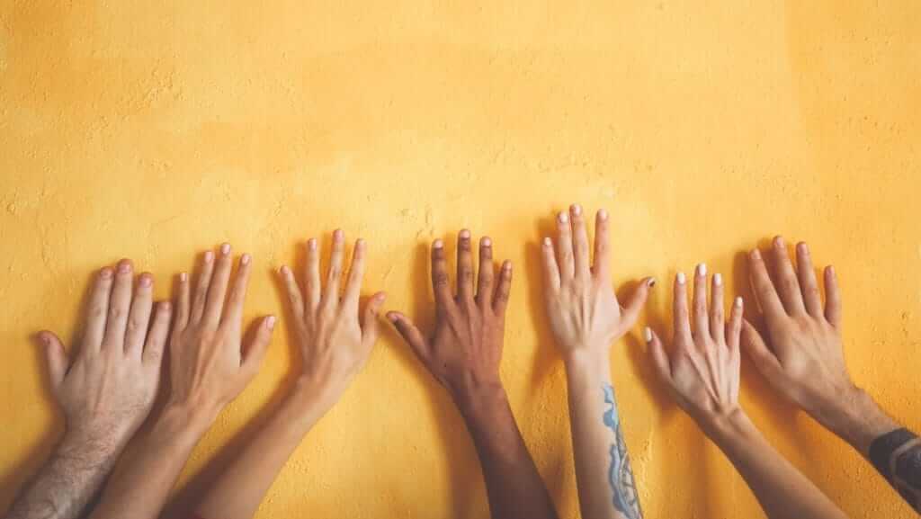 Several hands of diverse skin tones are outstretched against a bright yellow wall. The hands vary in size and some display unique tattoos, showcasing individuality and diversity. This vibrant image serves as a visual DEI tip, highlighting the beauty of inclusion and representation.