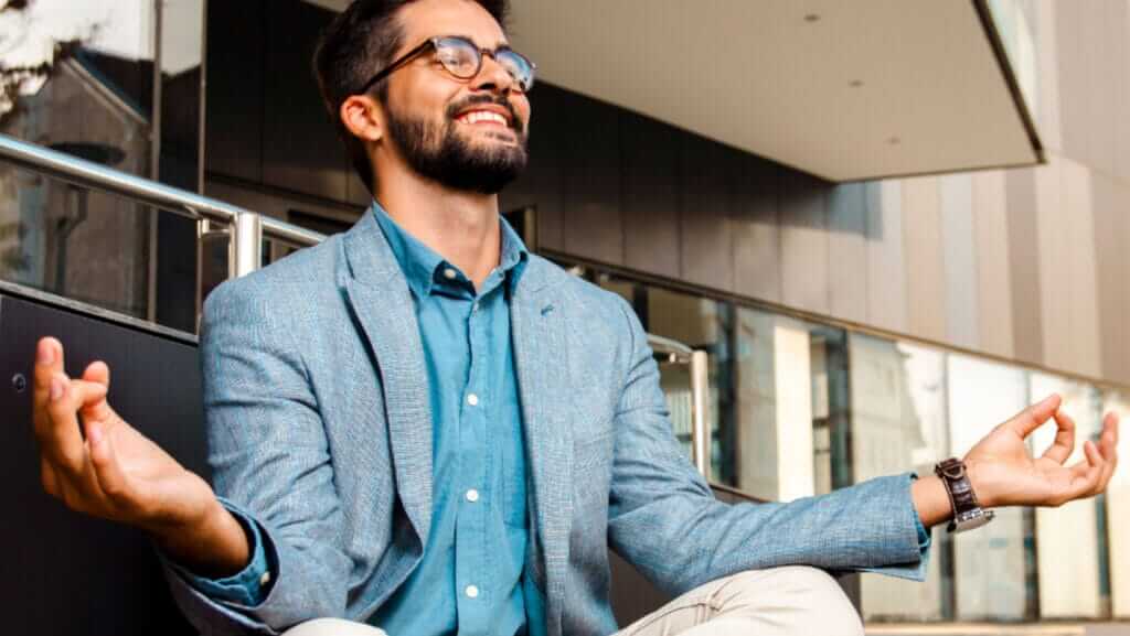 A bearded man wearing glasses, a blue shirt, and a light blue blazer sits cross-legged outdoors, smiling and meditating with his hands resting on his knees in a relaxed posture. Modern buildings and railings form the background as he reflects on the power of inspirational women.