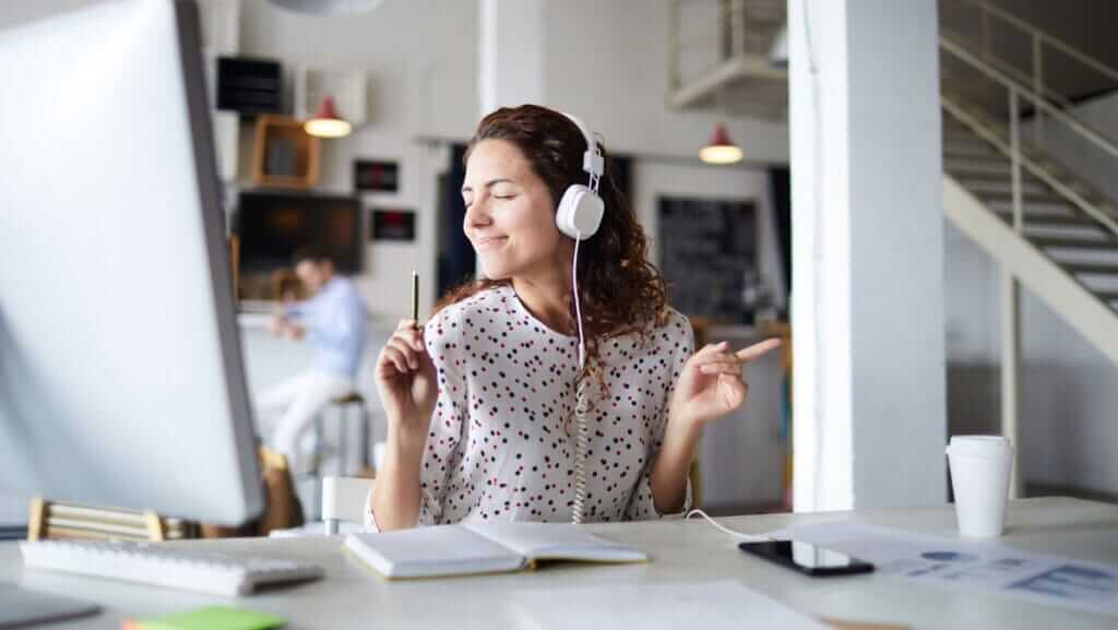 A woman with curly hair, wearing a white polka-dotted blouse, is sitting at a desk with a computer, headphones on, eyes closed, and smiling. She appears to be enjoying music while reviewing training courses, with one hand pointing and the other holding a pen. The background shows a modern office setting.