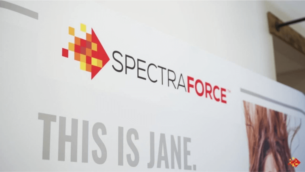 A display panel featuring the Spectraforce logo, which includes a pixelated arrow design in red and yellow, followed by the company name in black and red text. Below the logo, partially visible text reads "THIS IS JANE" on a light background, reminiscent of modern myths in storytelling.