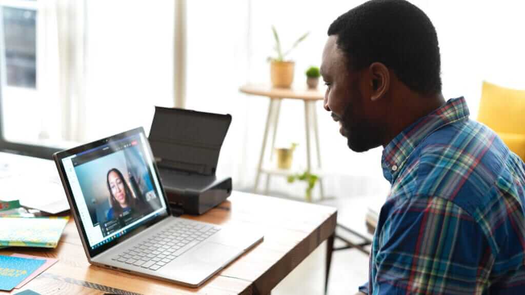 A person wearing a plaid shirt sits at a wooden desk while engaging in a video call on a laptop, likely discussing small business sustainability. The screen shows another individual participating in the call. Scattered papers, a printer, and a plant are visible in the background.