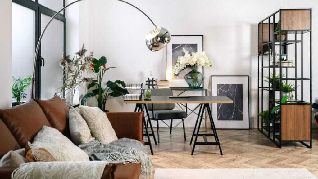 Modern living room and office space with a brown leather sofa adorned with cozy blankets and pillows. A sleek desk with a chair is set against a backdrop of potted office plants, framed artwork, and shelving units. A large arc floor lamp provides additional lighting.