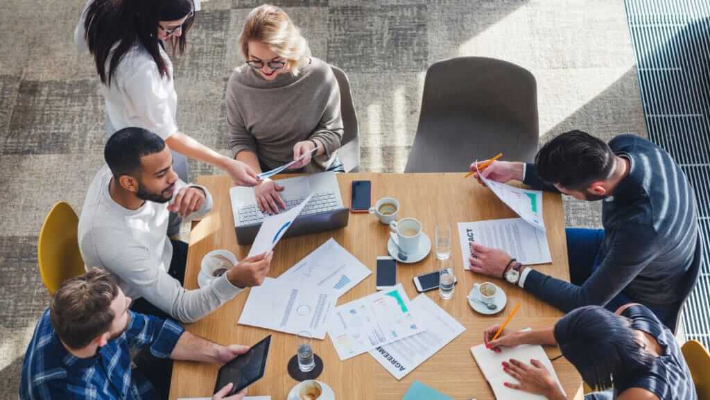 A diverse group of six people are gathered around a table in a collaborative meeting. They are engaged in discussion, reviewing documents, and working on laptops, likely planning upcoming training courses. The table is cluttered with papers, notebooks, coffee cups, and electronic devices.