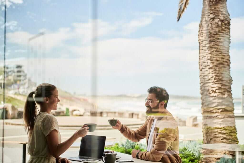 A man and woman are sitting at an outdoor café by the beach, enjoying coffee together. The woman, on the left, is holding a cup and smiling, while the bearded man, on the right, holds his cup up. A laptop is on the table with a job reference highlighted on its screen, and a palm tree is in the background.