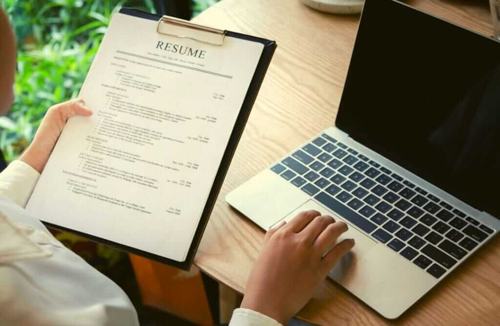 A person reviews a printed resume on a clipboard with their left hand while using a laptop placed on a wooden table with their right hand. The background shows a window with greenery outside, creating the perfect setting for evaluating both past experience and recent training courses.