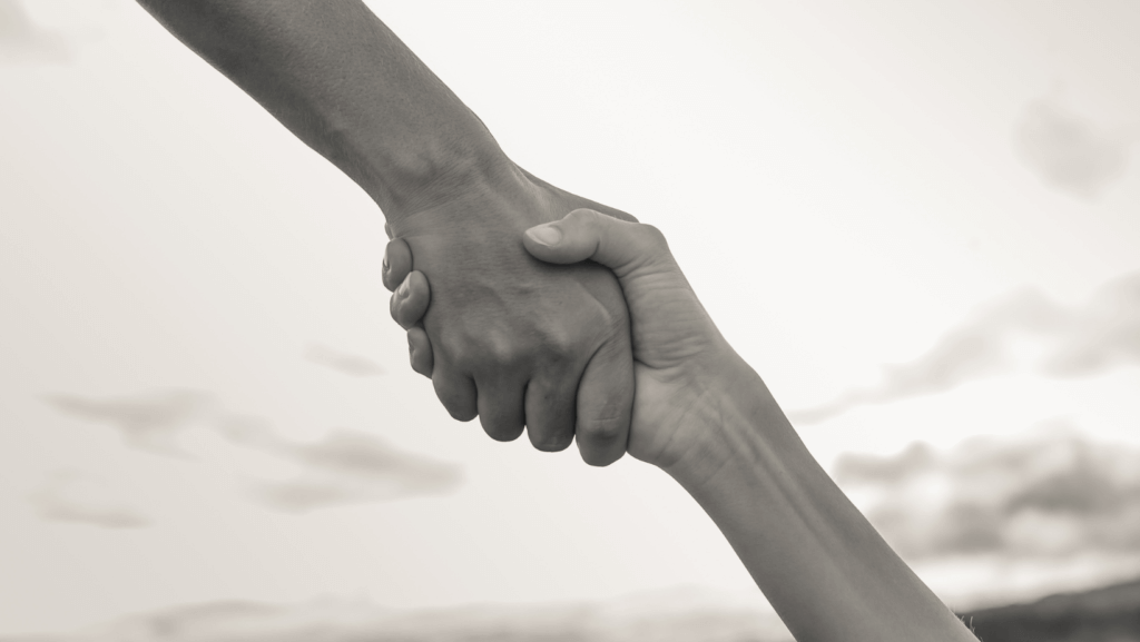 A close-up image shows two hands gripping each other firmly, symbolizing support and assistance. The background is blurred with soft, cloudy skies, emphasizing the focus on the hands. The image is in black and white, adding a timeless feel. The unity reflects a deep sense of sustainability and strength.