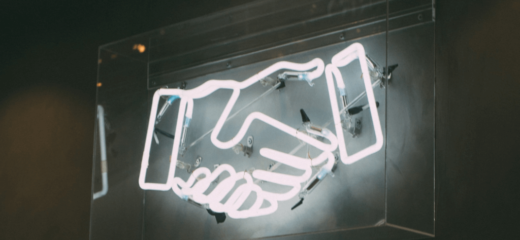 A glowing neon sign displaying the outline of two hands shaking, mounted on a metallic background. The sign is encased in a clear acrylic or glass frame.